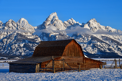 A wispy cloudbank starts to develop below the Tetons as the rising sun illuminates the famous and historic T.A. Moulton barn on a picture perfect March 2021 morning.