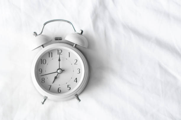 istock Alarm clock on white bad for wake up time with light from window, selective focus 1319157520