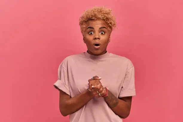 Photo of Dark-skinned woman with a very surprised expression stands on a pink background.