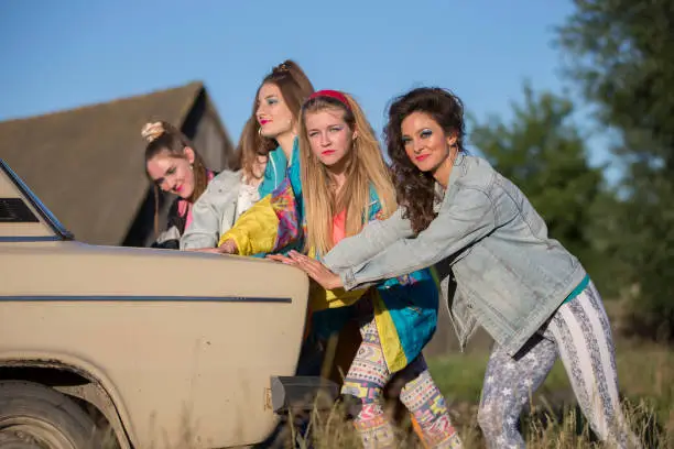 Photo of Young cheerful girls are pushing an old car. Women in the style of the 90s.