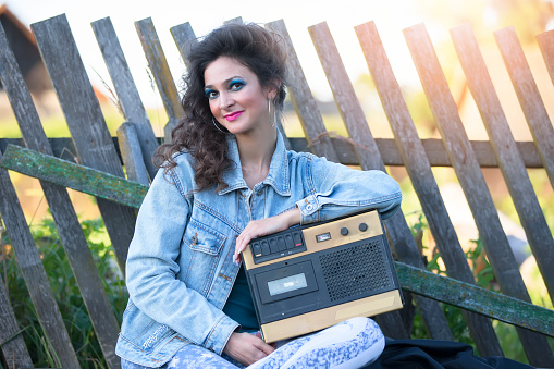 A beautiful country girl in bright clothes sits on a wooden bench with a cassette recorder. Woman in the style of the 90s.