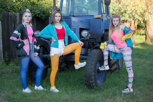 Three country girls dressed in the style of the 90s stand by the tractor.