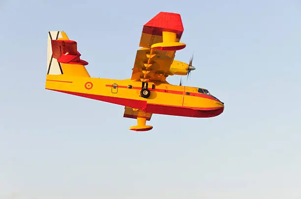 Close shot of flying firebrigade yellow and orange airplain, pale blue sky behind.