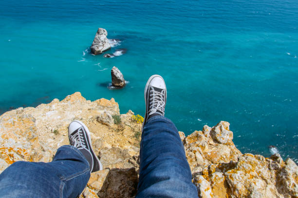 male legs in sneakers sitting on the edge of a high cliff and looking at the blue sea or ocean on a sunny summer day - hiking coastline waters edge sunny imagens e fotografias de stock