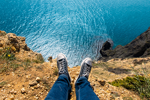 Male legs in sneakers sitting on the edge of a high cliff and looking at the blue sea or ocean on a sunny summer day