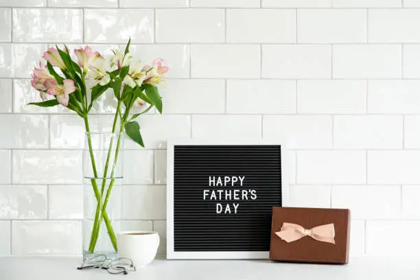 Happy Fathers Day concept. Letterboard with sign Happy Father's Day, gift box, boquet of flowers, coffee cup on table. Tile wall on background.