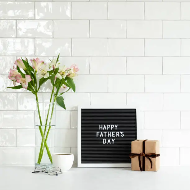 Happy Fathers Day concept. Letterboard with sign Happy Father's Day, gift box, boquet of flowers, coffee cup on table. Tile wall on background.