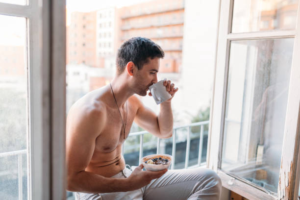 istock Shirtless young man sitting on an open window, having a breakfast and drinking coffee 1319182342