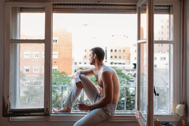 istock Shirtless young man sitting on an open window, having a breakfast and drinking coffee 1319182363