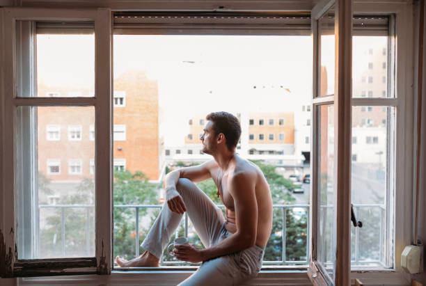 istock Shirtless young man sitting on an open window, having a breakfast and drinking coffee 1319182467