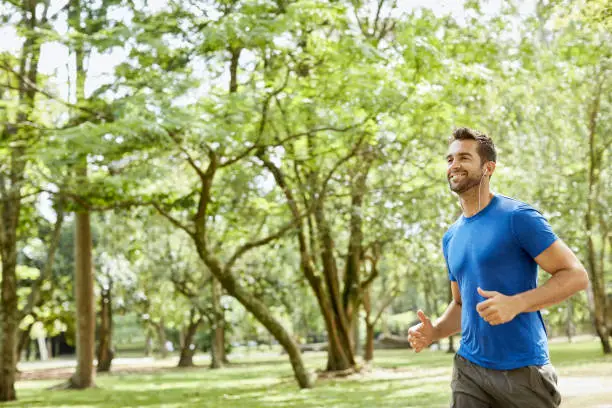 Smiling man with in-ear headphones jogging in park. Mid adult male is listening to music. He is wearing sports clothing.
