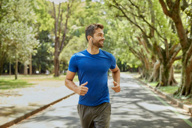 Mid adult man is listening to music in park Smiling male with in-ear headphones jogging in park. Mid adult man is listening to music during summer. He is wearing sports clothing. three quarter length stock pictures, royalty-free photos & images