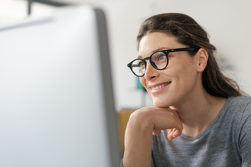 Woman with glasses working on a computer, she is smiling and staring at the screen