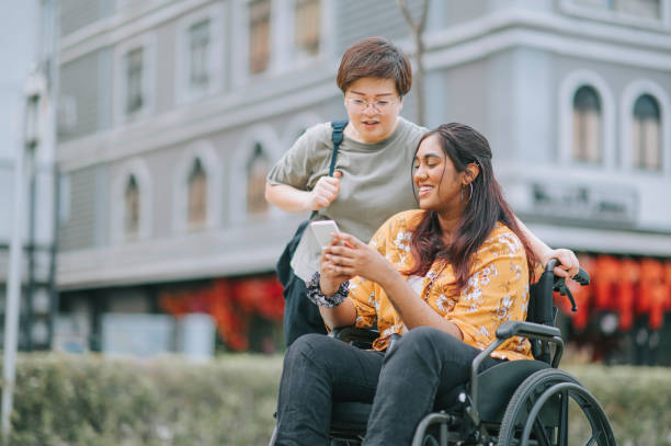 turista asiática india con silla de ruedas hablando con su amiga china en la acera de la ciudad - physical impairment wheelchair disabled accessibility fotografías e imágenes de stock