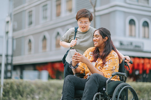 Tourist asian indian woman with wheelchair talking to her female chinese friend at sidewalk of the city