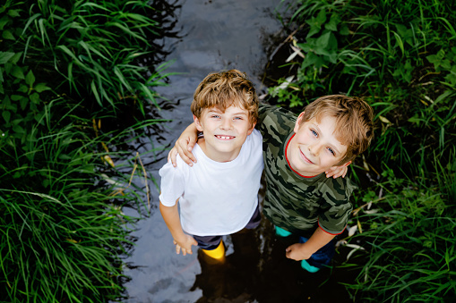 Two happy little school kids boys, funny siblings having fun together walking through water in river in gum rubber boots. Family portrait of healthy brothers and best friends.