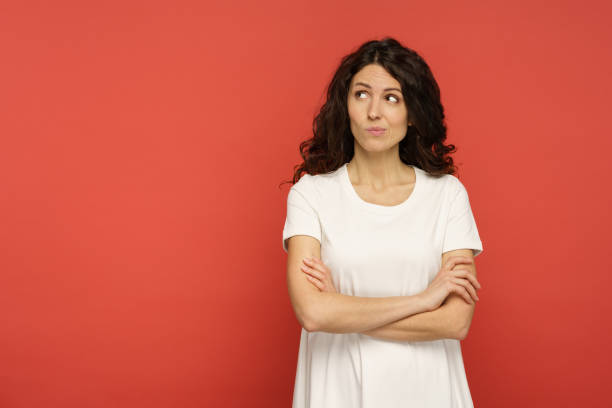 Sarcastic doubtful woman judgmental look with skeptical face aside to empty copy space over red wall Sarcastic doubtful young woman look with judgmental glance and skeptical face expression side to empty copy space over red wall background. Studio portrait of unsure suspicious female in white t-shirt sideways glance stock pictures, royalty-free photos & images