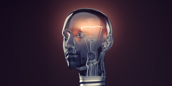 An upright glass lightbulb in the shape of a human head facing away from the camera in three quarter profile view with a glowing filament. The soft glow of the bulb lights the dark background creating a subtle halo. With copy space.