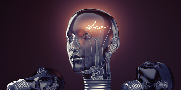 Three glass lightbulbs in the shape of a human head, two lying on the ground unlit, and one upright facing away from the camera in three quarter profile view with a glowing filament in the shape of the word 'idea'. The soft glow of the bulb lights the dark background creating a subtle halo. With copy space.