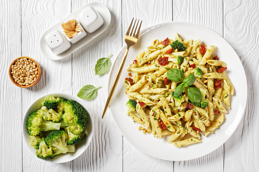 Broccoli pesto pasta with sundried tomatoes and pine nuts on a plate and ingredients on a  white wooden table, horizontal view from above, flat lay