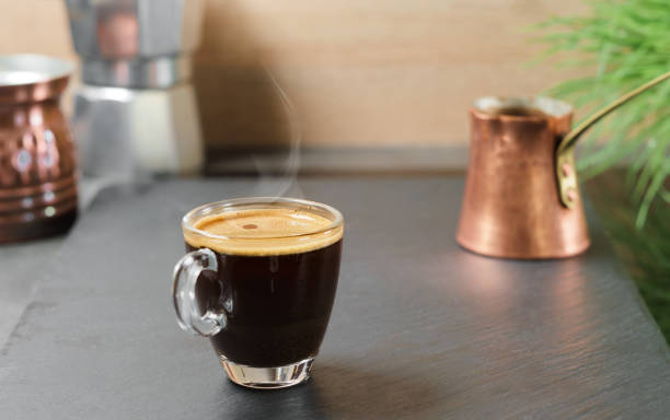 istock Coffee break. Fresh espresso steam rises in a glass goblet above the cup, selective focus. Strong aromatic coffee brewed in a Turk for breakfast, the idea of making coffee in traditional oriental way 1319215041