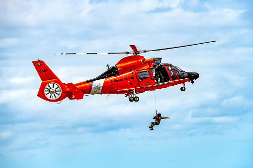 The Airbus MH-65 Dolphin helicopter is used by the US Coast Guard for search and rescue and show of force missions. This image shows the helicopter in a hover (stationary) state while lowering a rescue swimmer to the surface. The rescue swimmer is giving hand signals to the hoist operator.\nMelbourne, Florida\n)5/16/2021