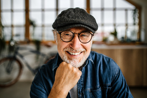 Handsome senior man with white beard, cap and eyeglasses, sitting in the livngroom looking at camera, smiling.