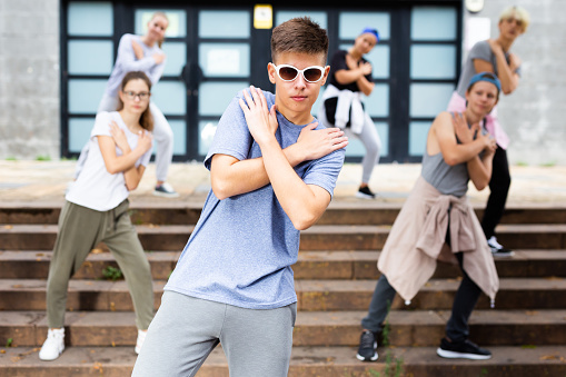 Positive teen boy dancing modern street dance outdoors with teenagers in background