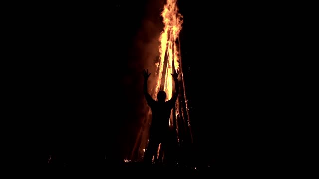 A man standing in the darkness in front of a big bonfire is holding his hands up and praying on it