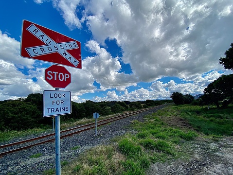 Horizontal landscape of red railway crossing stop sign with old country railway tracks with rocks green tree lined surrounds under a white cloudscape sky