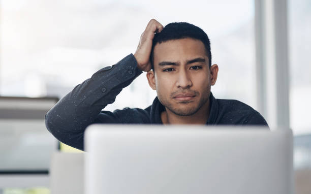 foto de un joven empresario guapo sentado solo en su oficina y con aspecto confuso mientras usaba su computadora portátil - scratching head fotografías e imágenes de stock