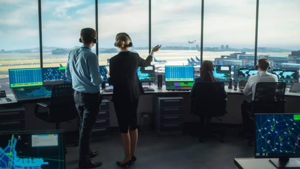 Female and Male Air Traffic Controllers with Headsets Talk in Airport Tower. Office Room is Full of Desktop Computer Displays with Navigation Screens, Airplane Departure and Arrival Data for the Team. Female and Male Air Traffic Controllers with Headsets Talk in Airport Tower. Office Room is Full of Desktop Computer Displays with Navigation Screens, Airplane Departure and Arrival Data for the Team. air traffic control tower stock pictures, royalty-free photos & images