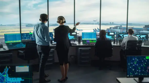 Photo of Female and Male Air Traffic Controllers with Headsets Talk in Airport Tower. Office Room is Full of Desktop Computer Displays with Navigation Screens, Airplane Departure and Arrival Data for the Team.