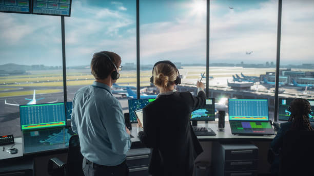 Female and Male Air Traffic Controllers with Headsets Talk in Airport Tower. Office Room is Full of Desktop Computer Displays with Navigation Screens, Airplane Departure and Arrival Data for the Team. Female and Male Air Traffic Controllers with Headsets Talk in Airport Tower. Office Room is Full of Desktop Computer Displays with Navigation Screens, Airplane Departure and Arrival Data for the Team. atc stock pictures, royalty-free photos & images