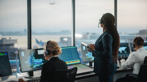空港タワーにタブレットを保持する黒い女性航空管制官。オフィスルームは、ナビゲーション画面、飛行機の出発と到着のデータチームのデスクトップコンピュータディスプレイでいっぱい� - air traffic control tower ストックフォトと画像