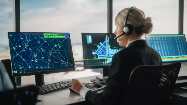 Photo of Female Air Traffic Controller with Headset Talk on a Call in Airport Tower. Office Room is Full of Desktop Computer Displays with Navigation Screens, Airplane Flight Radar Data for the Team.