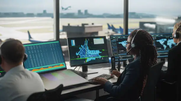 Photo of Female Air Traffic Controller with Headset Talk on a Call in Airport Tower. Office Room is Full of Desktop Computer Displays with Navigation Screens, Airplane Flight Radar Data for the Team.