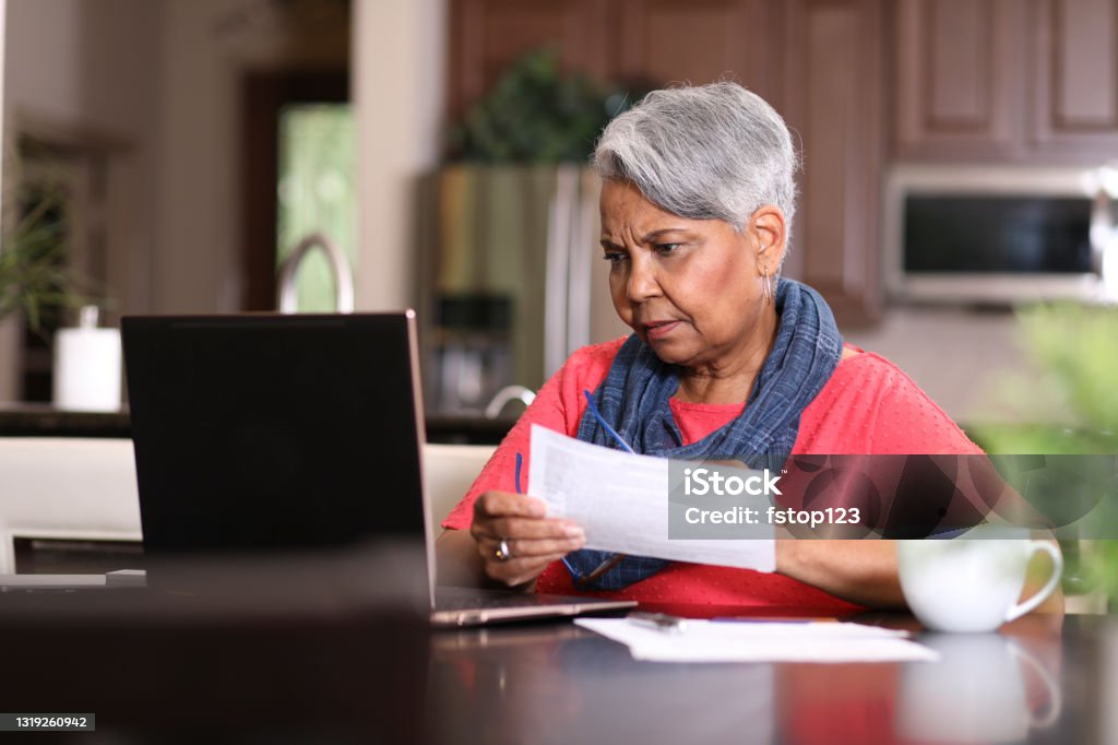 Senior adult woman frustrated paying bills at home. African descent, senior adult woman at home sitting at kitchen table paying bills or online banking using laptop computer. She is frustrated at the overdue charges. Paperwork and coffee cup on table. Senior Adult Stock Photo