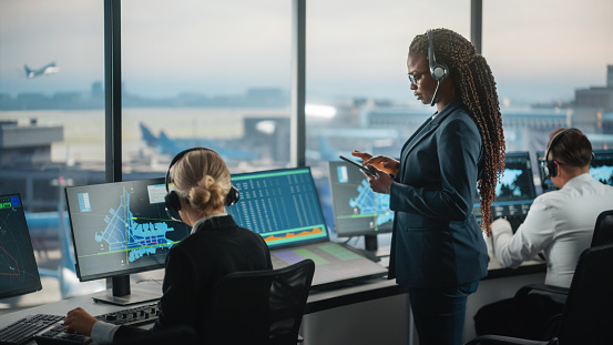 Black Female Air Traffic Controller Holding Tablet in Airport Tower. Office Room is Full of Desktop Computer Displays with Navigation Screens, Airplane Departure and Arrival Data for the Team.