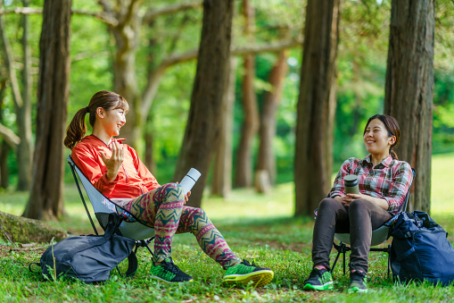 Two female hikers taking break from walking and sitting and drinking water while enjoying talking with each other in nature