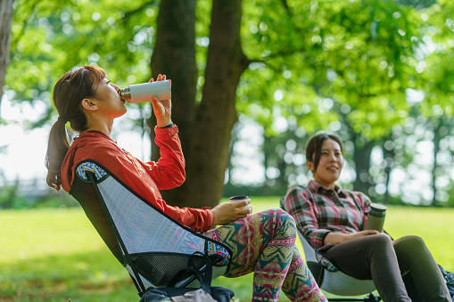 Two female hikers are taking a break from walking and sitting and drinking water while enjoying talking with each other in nature.