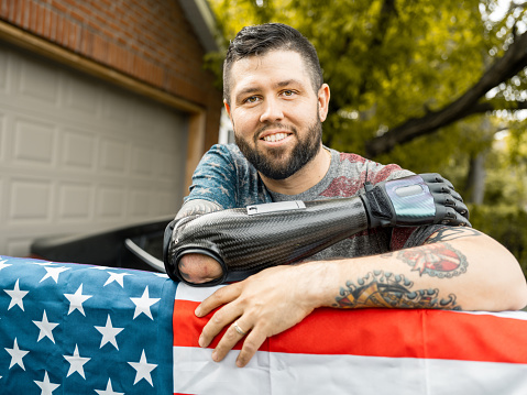 Caucasian Man with disability- prosthetic lower arm posing with American flag in his backyard. He is wearing casual outfit with American flag motif  t-shirt. Exterior of urban backyard of private home.