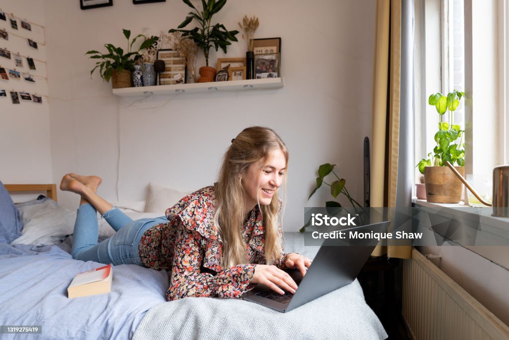 A young woman lying on bed Young university student following lecture on her laptop on the bed College Dorm Stock Photo