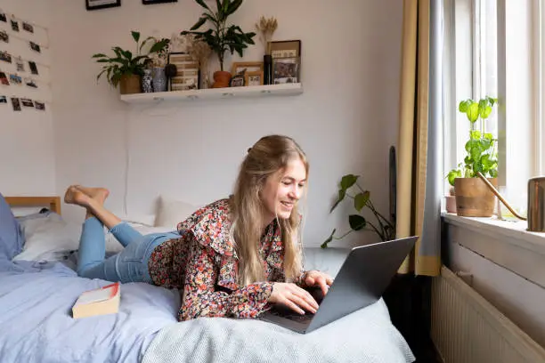 Young university student following lecture on her laptop on the bed