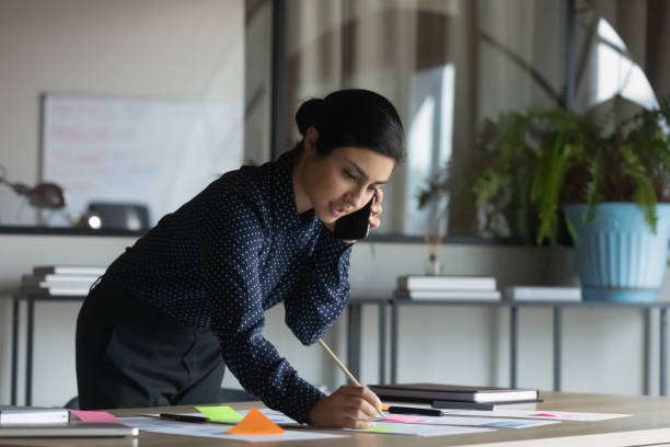 istock Young Indian female employee talk on cellphone work with paperwork 1319284379