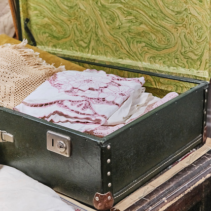 An antique white handmade tablecloth with cutwork embroidery lies in an old suitcase.
