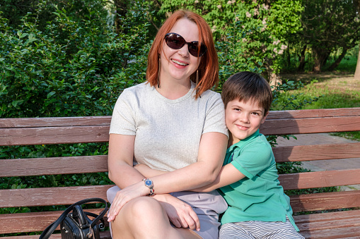 Young woman with red hair and boy are sitting in park on bench and smile. Son hugs his mother gently. Happy parenting concept, relationship with children. Attachment of young children to their mother.