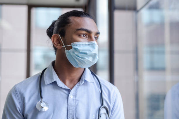 Global lockdowns during the COVID-19 pandemic Portrait of male doctor of Indian descent looking out the window. Wearing a protective face mask and stethoscope around his neck. Modern hospital setting. self sacrifice stock pictures, royalty-free photos & images