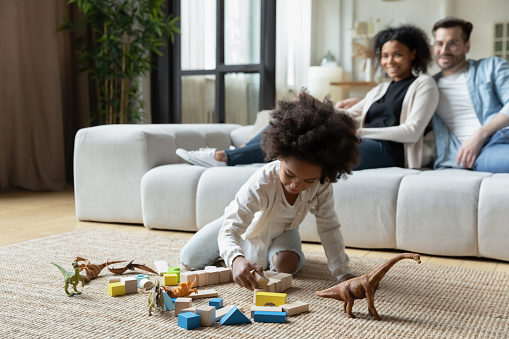 Small African American little girl child play with toy animals in living room, happy multiethnic mom and dad relax on sofa. Loving multiracial young family with daughter rest at home. Rental concept.