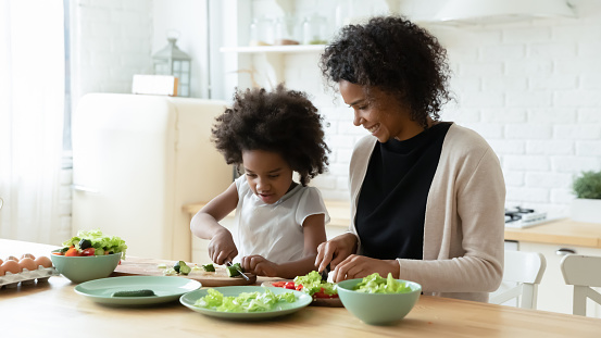 Smiling loving African American mother and little daughter prepare healthy delicious salad in kitchen. Happy caring biracial mom and small ethnic girl child cook together at home. Family concept.